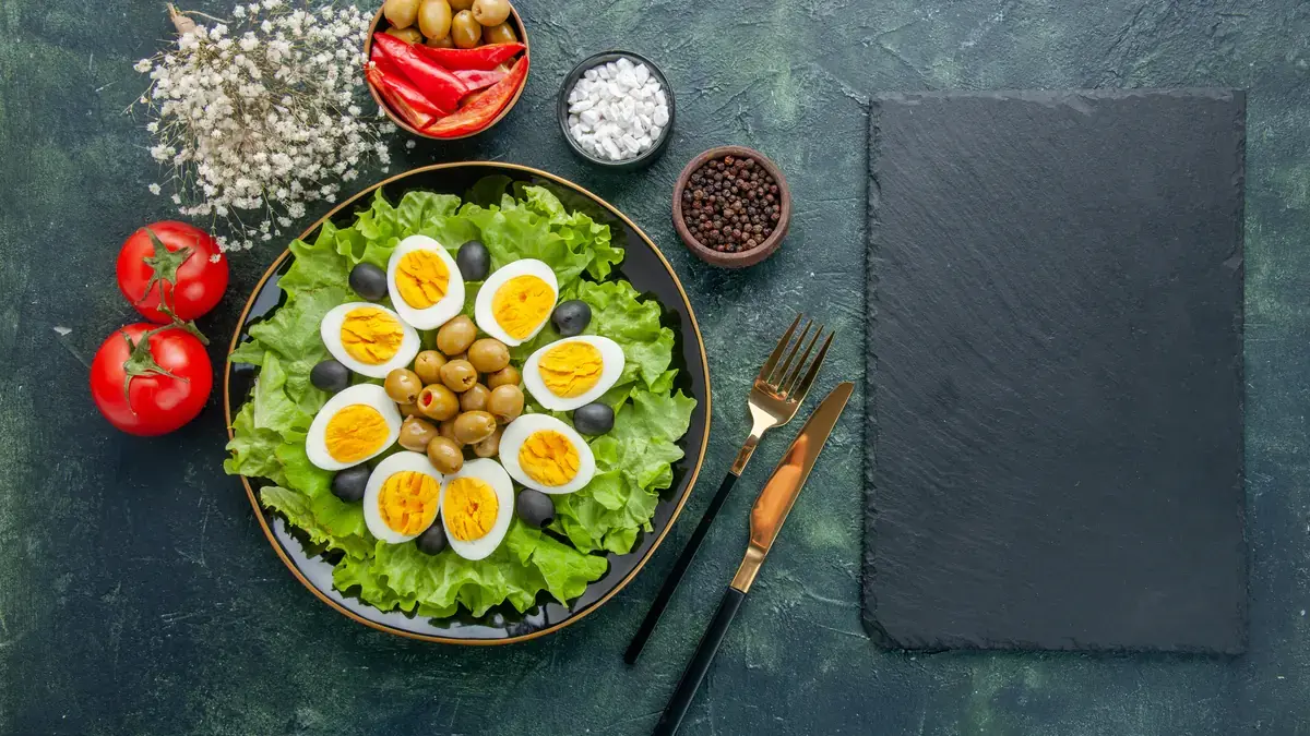 Sliced boiled eggs on a green salad with olives, tomatoes and spices on a dark table