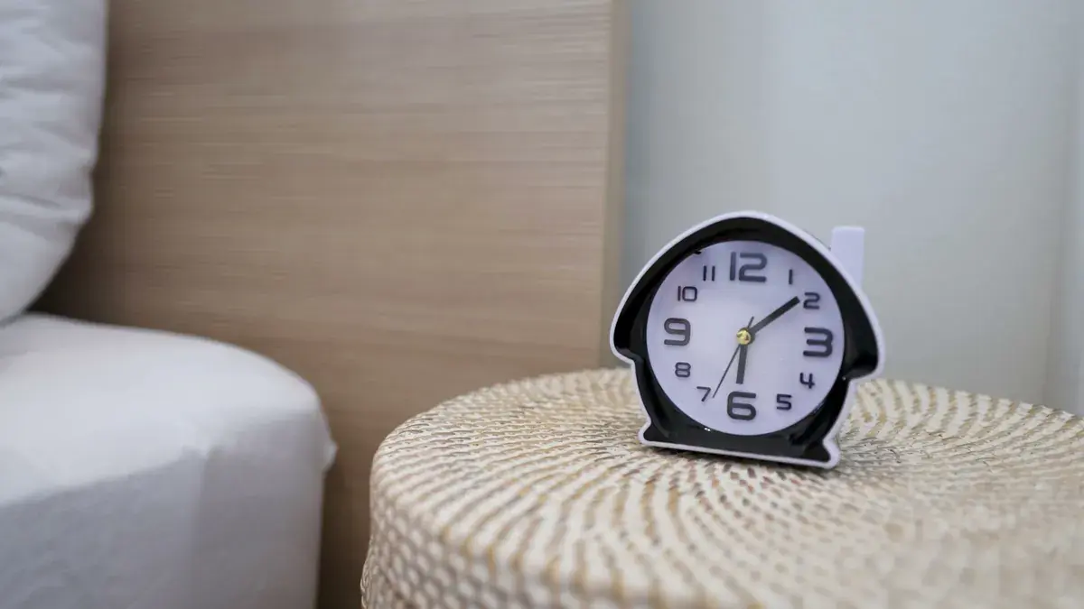 An alarm clock on a woven bedside table next to a bed